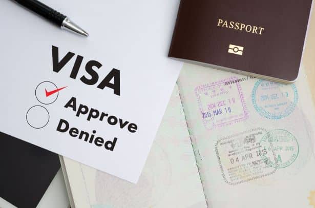 Obraz przedstawia formularz zatwierdzenia wizy z zaznaczoną opcją „Approve” (Zatwierdzono) i pustym okręgiem przy opcji „Denied” (Odrzucono). Na obrazku widoczne są również długopis, paszport oraz strona z paszportu z kilkoma pieczątkami wizowymi, datowanymi na 20 grudnia 2013, 10 marca 2015 i 4 kwietnia 2015.