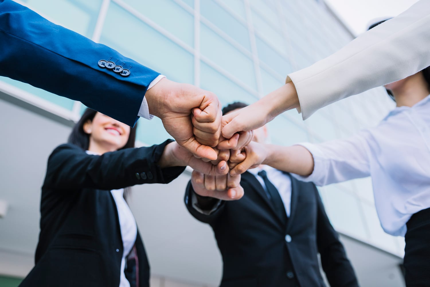 Obraz przedstawia grupę ludzi ubranych w stroje biznesowe, stojących w kręgu i wykonujących gest fist bump. W tle znajduje się nowoczesny budynek z dużymi szklanymi oknami.