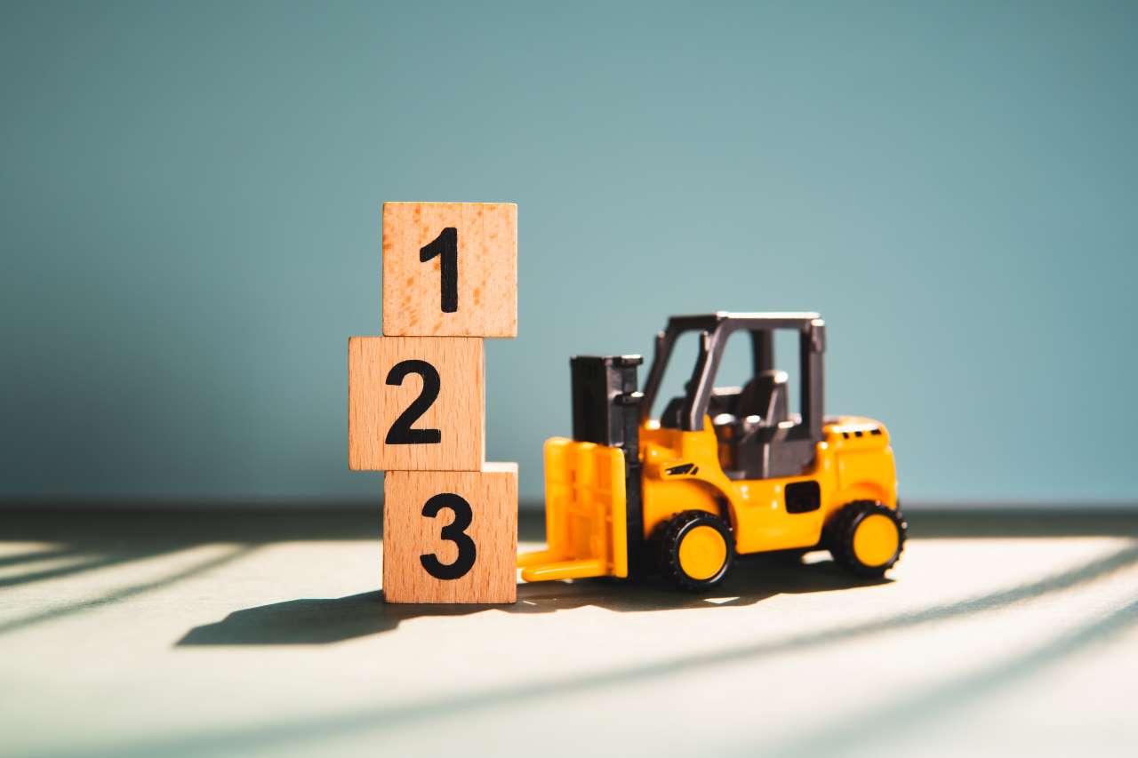 JThe picture shows a small yellow toy forklift next to a pile of three wooden blocks. The blocks are arranged vertically, and each of them has a number on it. From top to bottom, these are the numbers 1, 2, and 3. The forklift looks as if it is in the process of lifting or stacking blocks.