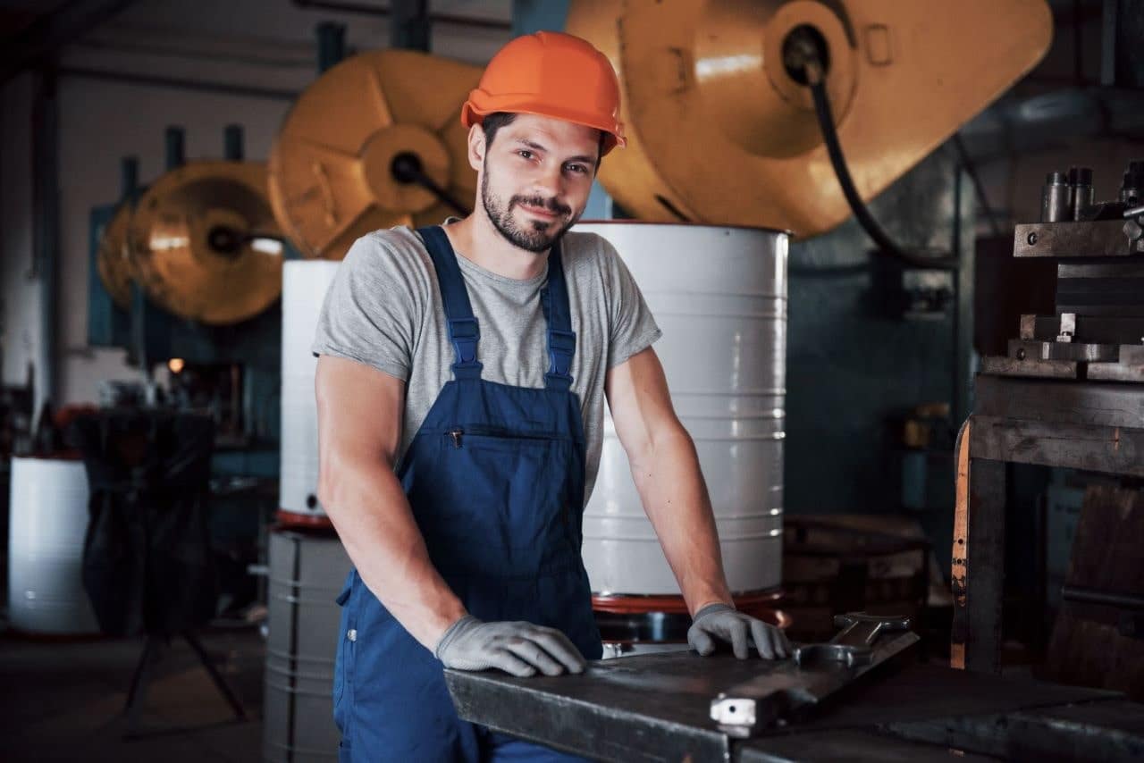 Obraz przedstawia osobę w pomarańczowym kasku ochronnym, szarym t-shircie i niebieskim kombinezonie, stojącą w produkcyjnym otoczeniu. Osoba opiera się rękami w rękawicach na metalowej powierzchni. W tle widać duże maszyny przemysłowe i urządzenia, w tym duże cylindryczne zbiorniki i maszyny z okrągłymi komponentami..