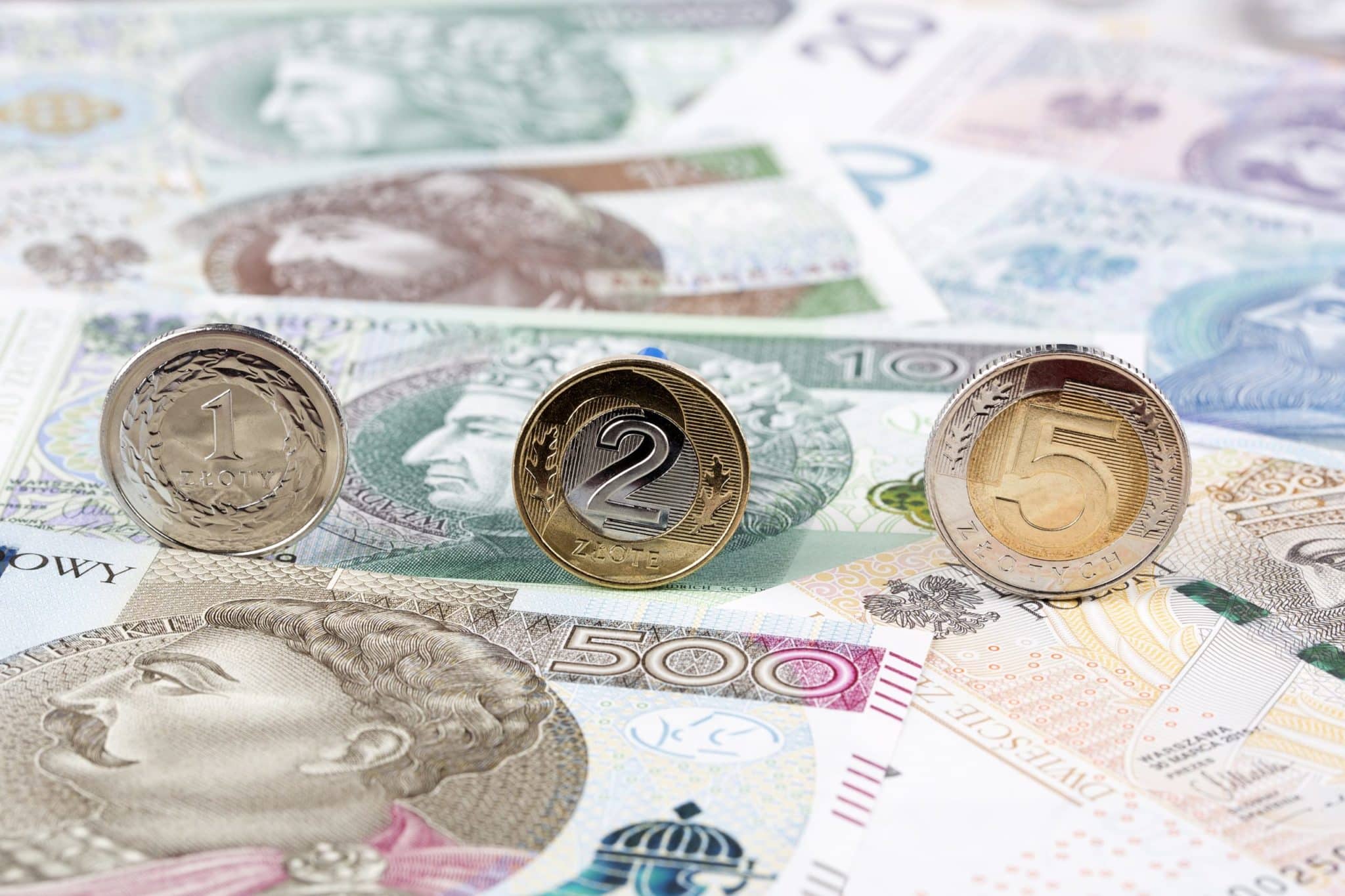 Na zdjęciu widzimy polskie monety i banknoty. W centrum kompozycji znajdują się monety o nominałach 1 zł, 2 zł i 5 zł, ustawione pionowo na tle rozłożonych banknotów. Banknoty mają różne nominały, w tym widoczny banknot o wartości 500 zł.