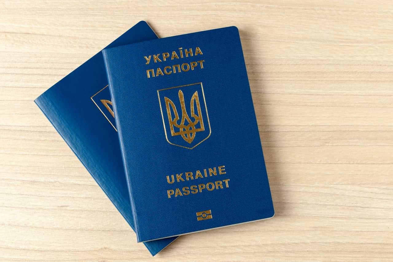 Na zdjęciu widoczne są dwa ukraińskie paszporty biometryczne, ułożone na jasnym, drewnianym blacie. Paszporty mają niebieską okładkę z wyraźnym złotym nadrukiem zawierającym napis "Україна Паспорт" (Ukraina Paszport) oraz "Ukraine Passport".