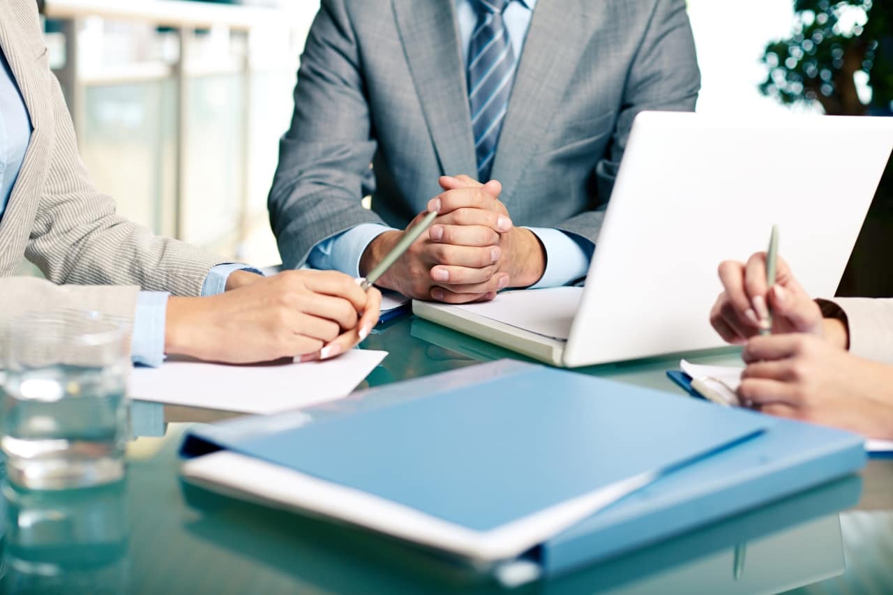 Na zdjęciu widoczne są trzy osoby w profesjonalnym otoczeniu, prawdopodobnie podczas spotkania biznesowego. Siedzą one przy stole, na którym znajdują się dokumenty, laptop oraz szklanka z wodą. Dwie osoby trzymają długopisy, sugerując, że mogą robić notatki lub omawiać dokumenty. Uczestnicy są ubrani w biznesowe stroje