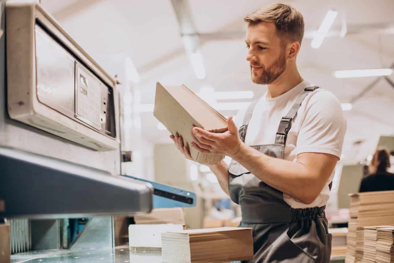 Obraz przedstawia mężczyznę w środowisku pracy, prawdopodobnie w fabryce lub warsztacie, trzymającą stos kartonu lub papieru. Mężczyzna nosi biały koszulę i szare spodnie. W tle znajduje się maszyna lub urządzenie, które wydaje się być związane z zadaniami wykonanymi przez osobę.