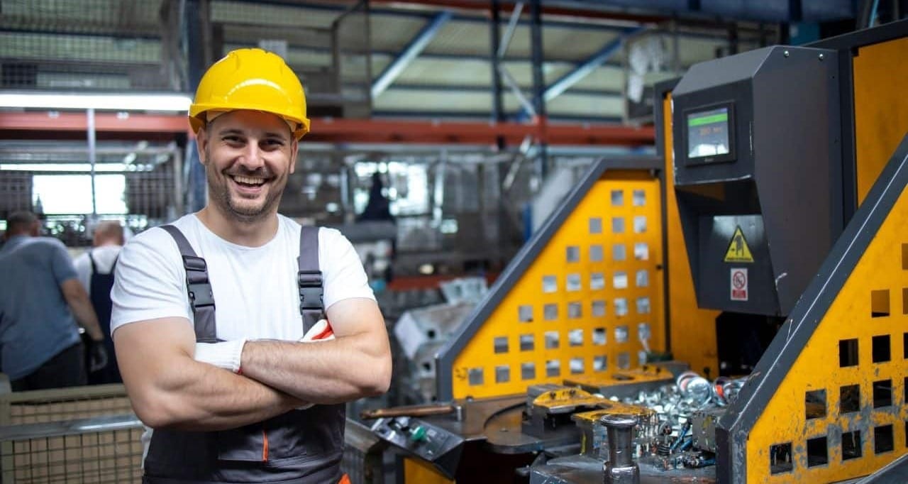 The image shows a smiling man wearing a yellow safety helmet and a white shirt with dark overalls, standing with his arms folded in an industrial setting.