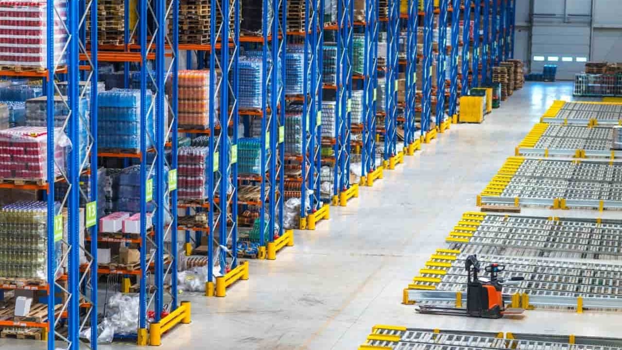 The painting shows the interior of a large warehouse equipped with tall, blue metal shelves filled with a variety of goods and products. The shelves are arranged in rows, and the items are stored on pallets. There are yellow protective barriers at the bottom of the shelves. On the right side of the painting are conveyor belts and a small forklift or pallet truck.