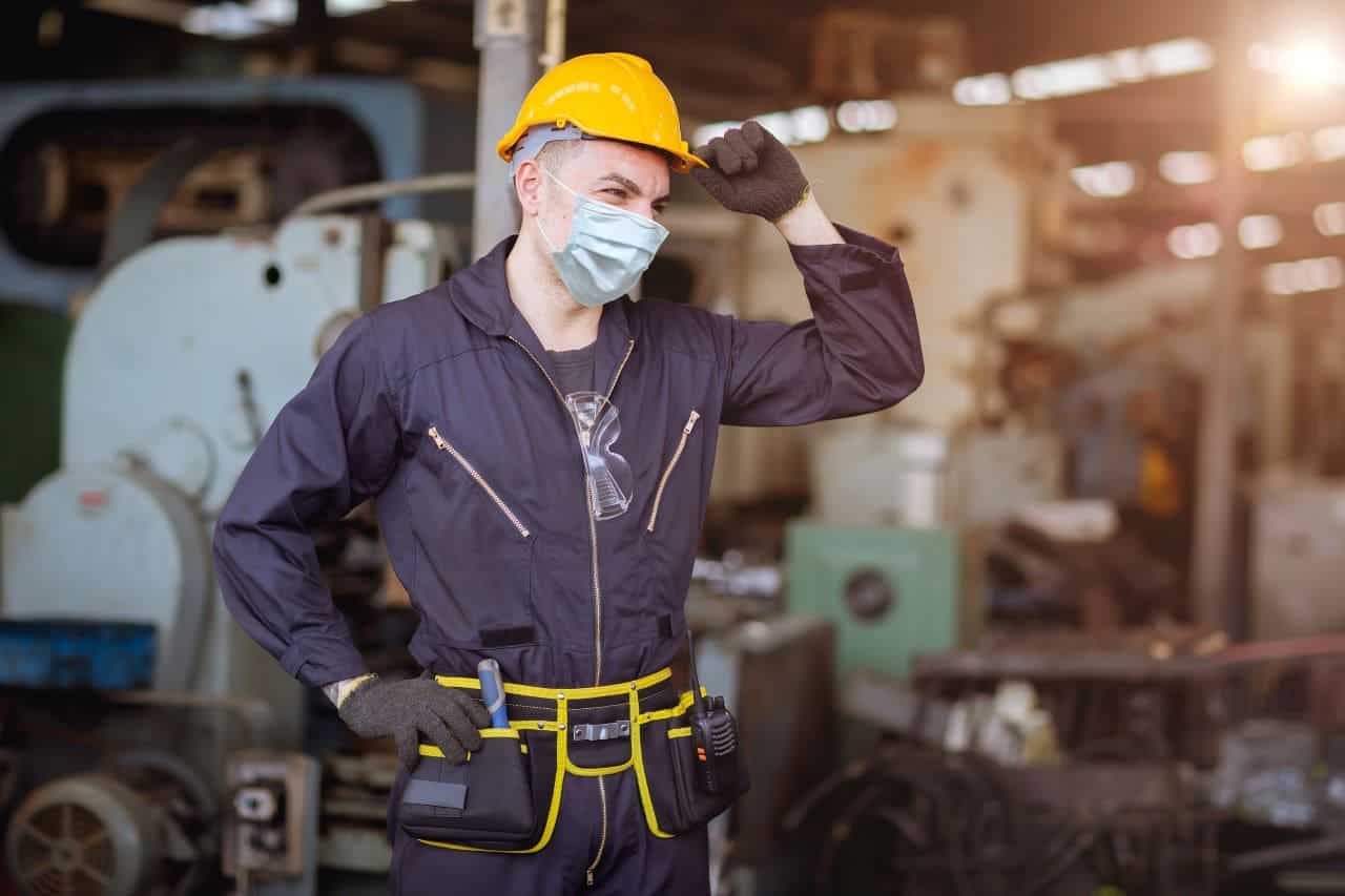 The photo shows a man wearing a dark blue work suit with yellow accents and a yellow hard hat. The man is in a factory surrounded by machinery and equipment. He wears a tool kit on his belt, including a walkie-talkie. He holds his hard hat with one hand and rests the other on his hip.