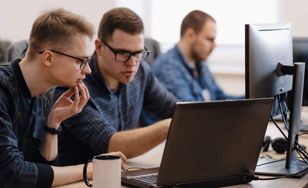 The photo shows three men working together in an office setting. Two of them are focused on a laptop screen, presumably discussing some topic, while the third man is working at a desktop computer.
