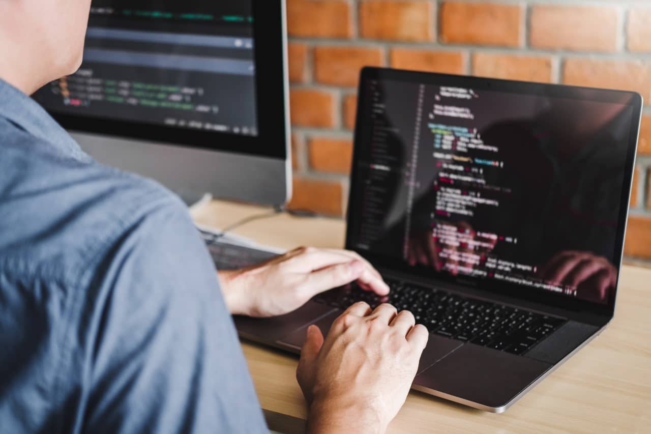 The photo shows a person working on a laptop with programming code displayed on the screen. The person is typing on the keyboard, and in the background is a second monitor, also displaying code. The room has an industrial feel, with a brick wall in the background, suggesting a studio or office.