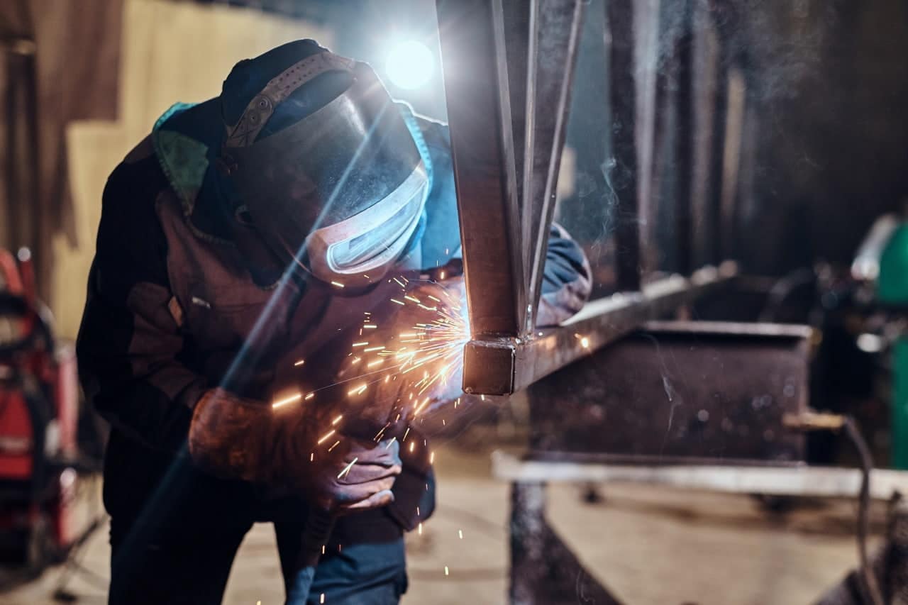 The photo shows a person wearing protective gear, including a welding visor and gloves, welding a metal structure. Sparks are rising from the welding site, indicating active work. In the background, there are elements of industrial equipment and tools, suggesting a workshop or production hall.