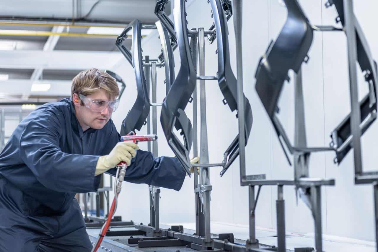 The photo shows a man wearing a blue protective suit and gloves using a spray gun to apply a coating or paint to metal frames. These frames are mounted on a stand in an industrial or manufacturing environment. The process is likely part of a production line where metal parts are prepared for further assembly or finishing.