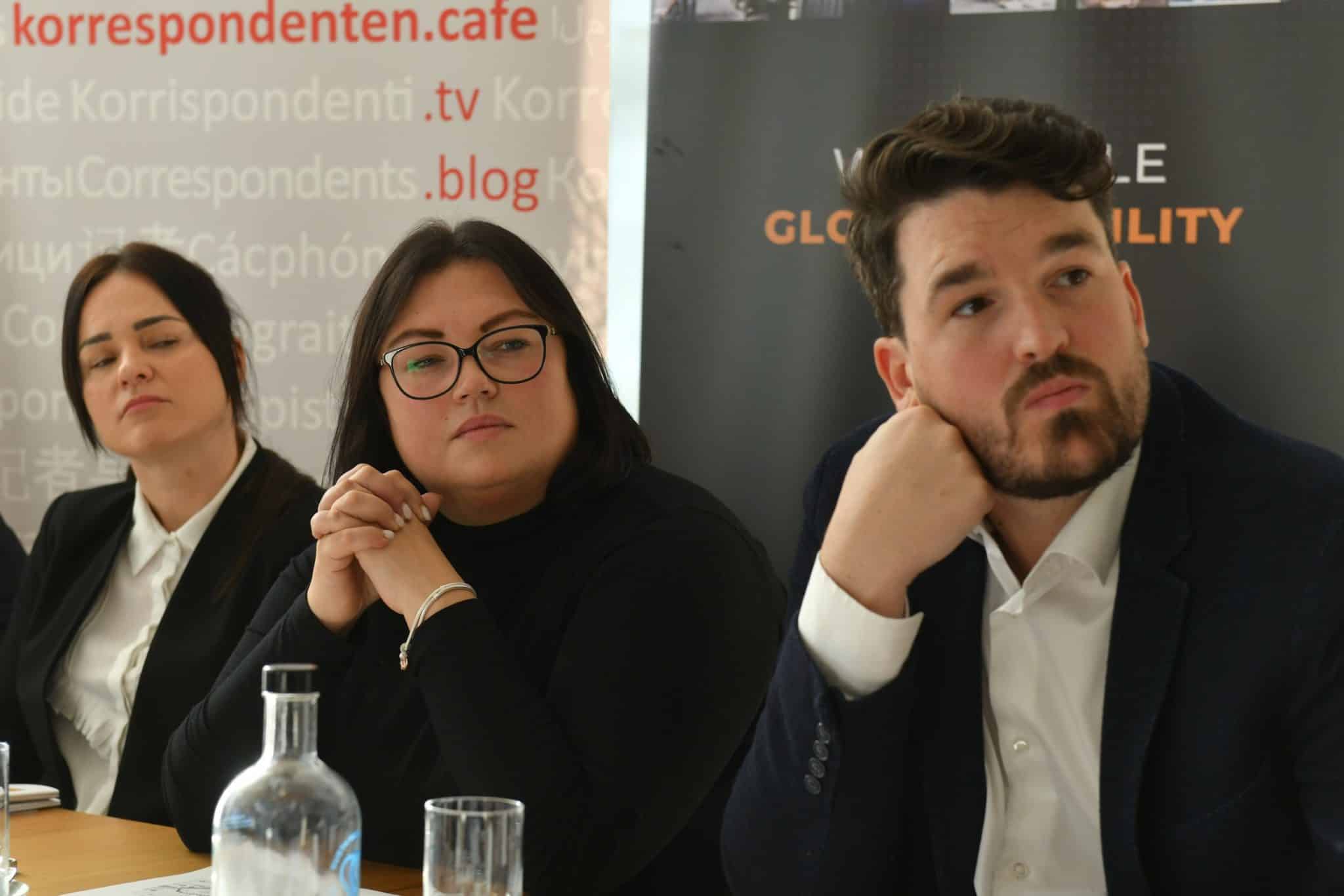 The photo shows three people sitting at a table, presumably attending a conference. In the background is a banner with text in multiple languages, including names such as “korrespondenten.cafe,” “Korrespondenti.tv,” and “Korrespondents.blog.” There is also a black banner with the words “GLOBAL” and “QUALITY” partially visible. On the table are a bottle of water, a glass, and several documents.