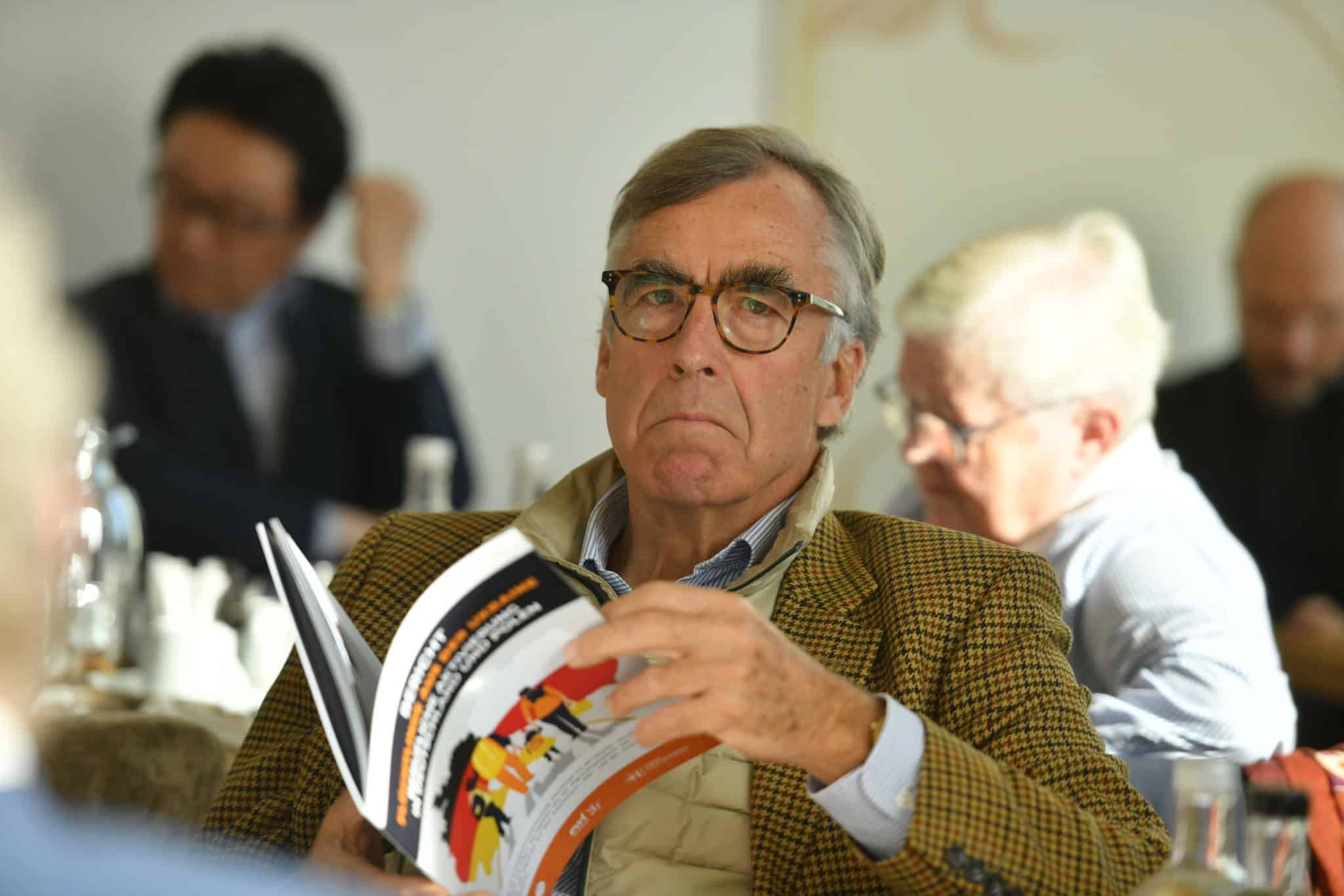 The image shows a man holding and reading a book or magazine. The cover of the publication is colorful and contains illustrations of characters and text. The man is wearing a brown, plaid blazer over a light shirt. In the background are other people sitting at tables, and on the table in front of him are glass bottles and cups.
