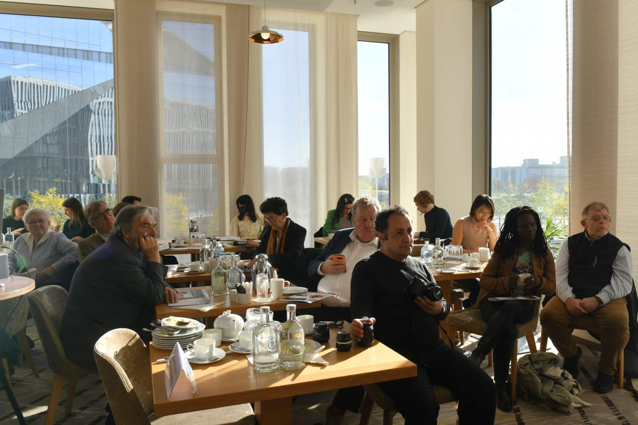 The photo shows a group of people sitting at tables in a well-lit room with large windows. There are various items on the tables, including bottles of water, cups, plates, and documents. Some of the participants are having conversations, while others appear to be listening or working on something. Large windows are visible in the background, giving a view of the building outside.
