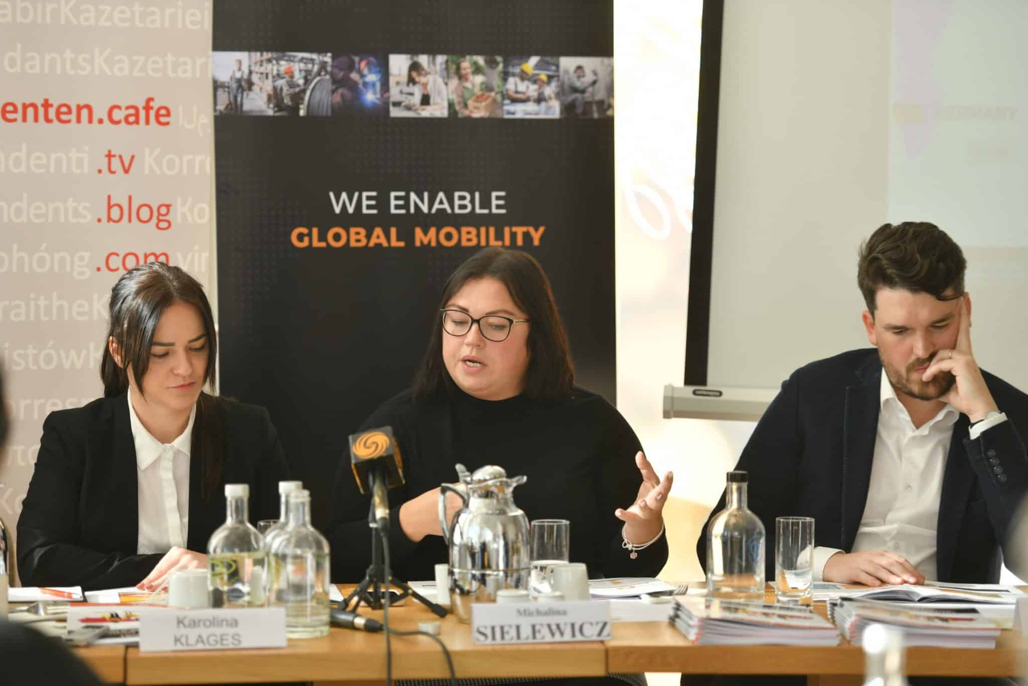 The photo shows three people sitting at a table during a conference or meeting. In front of each participant are name tags: Karolina Klages, Michalina Sielewicz and a third, unidentified person. In the background is a banner with the words "WE ENABLE GLOBAL MOBILITY" and illustrations of various activities.
