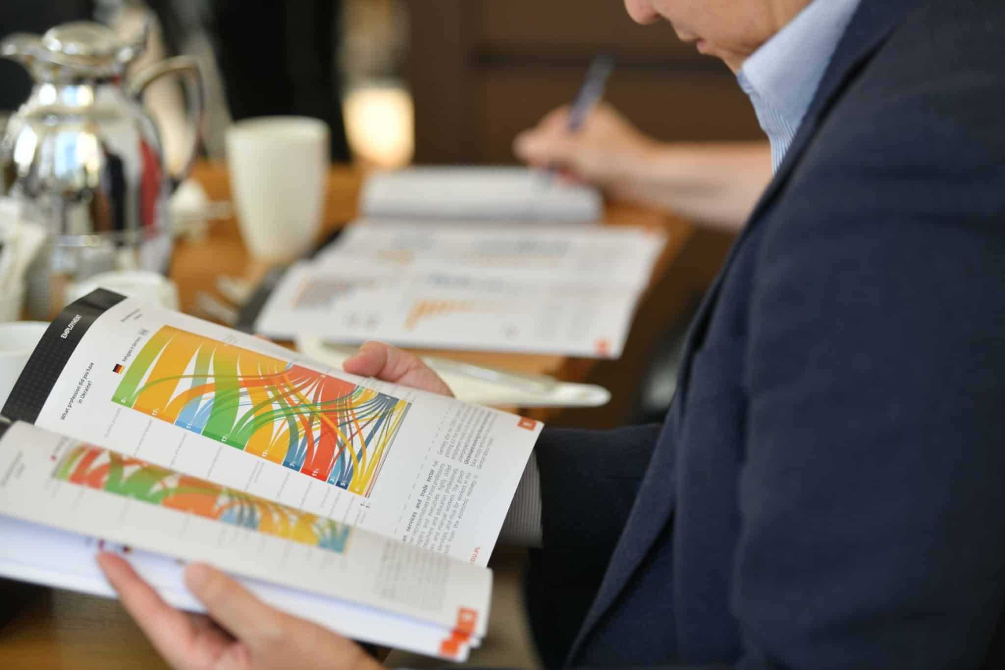 The photo shows a man in a suit reading a brochure or magazine containing colorful charts and graphs. The man is seated at a table on which there is a coffee pot, a cup, and other documents or brochures.
