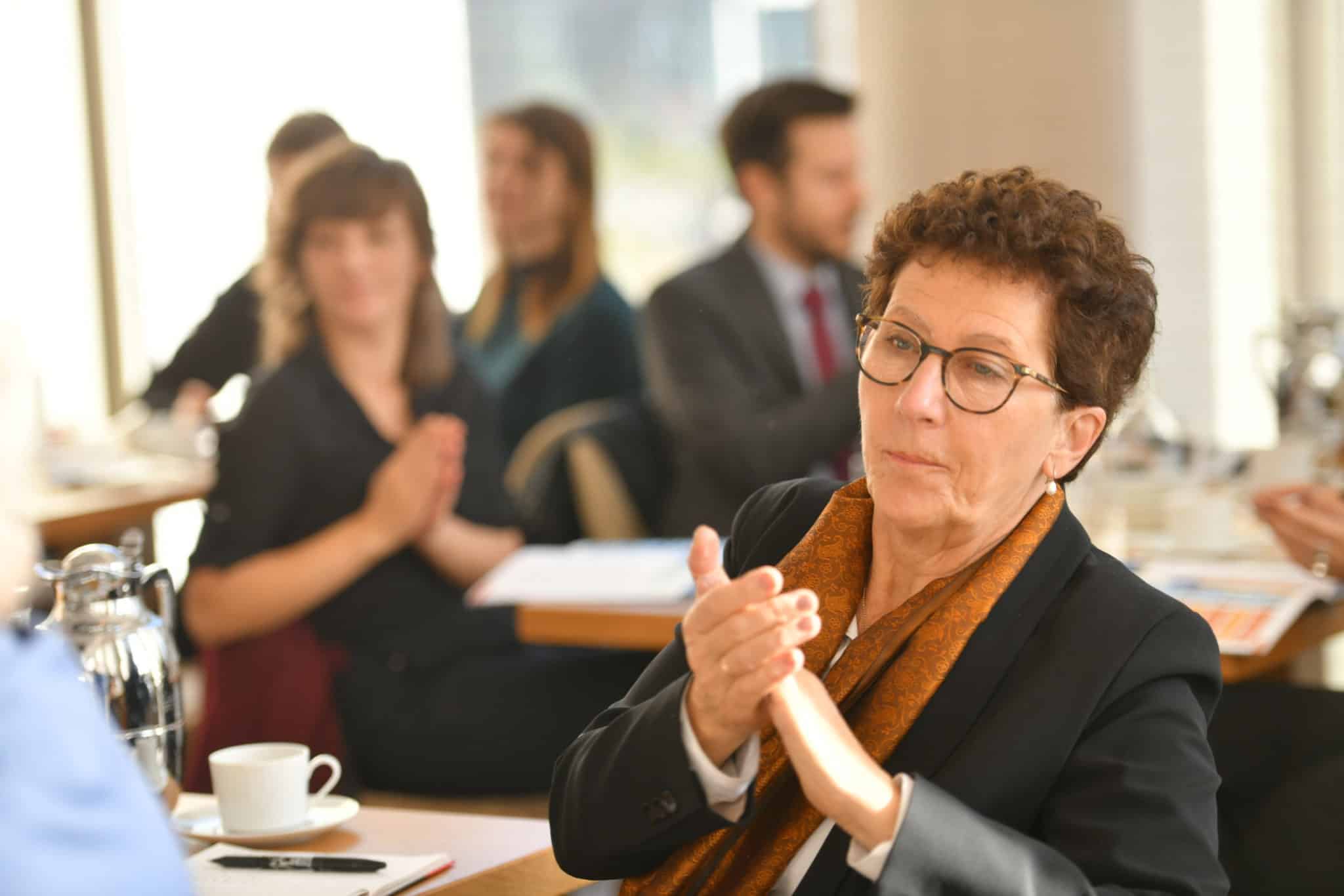 The photo shows a group of people sitting at tables in a setting resembling a conference or formal meeting. The participants are clapping, suggesting that they are appreciating or celebrating something. In the foreground is a woman with short hair. She is wearing a black blazer and a brown scarf.
