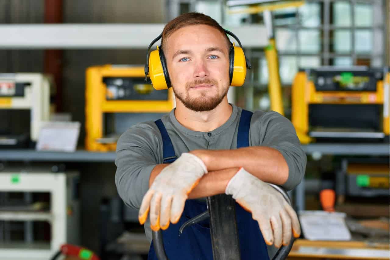 The photo shows a smiling man in a workshop environment. He is wearing grey long sleeves, blue dungarees and yellow ear protection. He is wearing white gloves with orange tips. He is leaning against a pallet jack. In the background are yellow and black machines, probably carpentry tools such as planers or sanders.