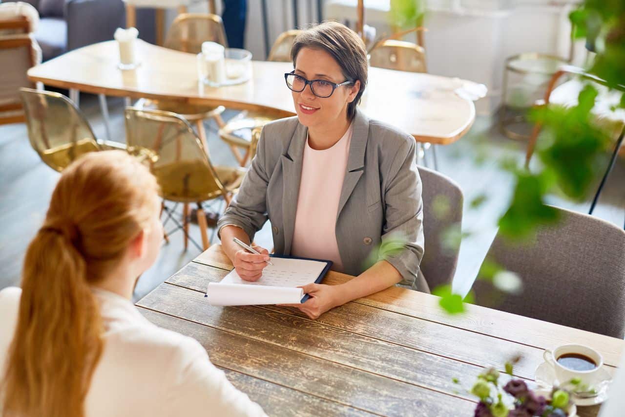 Das Foto zeigt zwei Frauen, die in einem hellen, modernen Ambiente an einem Tisch sitzen, möglicherweise in einem Café oder Restaurant. Eine Frau, die von hinten zu sehen ist, hat die Haare zusammengebunden und trägt helle Kleidung. Die andere Frau, die ihr gegenüber sitzt, hat kurze Haare, trägt eine graue Jacke und hält einen Stift und ein Notizbuch in der Hand, in das sie etwas schreibt. Auf dem Tisch steht eine Tasse mit einem Getränk und anderen Gegenständen. Im Hintergrund sind weitere Tische und Stühle zu sehen.