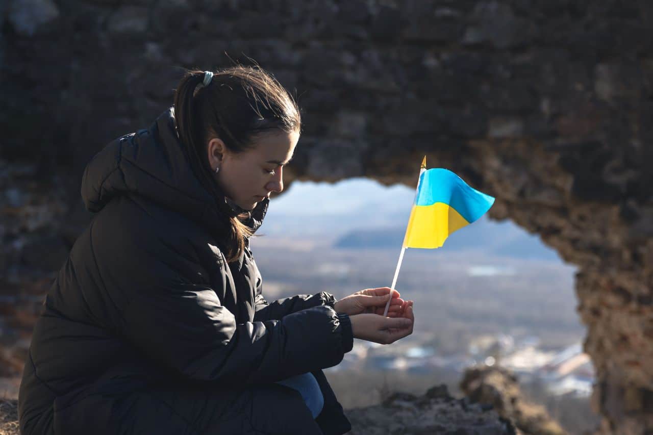 Das Gemälde zeigt eine Frau, die im Freien sitzt und eine kleine ukrainische Flagge in der Hand hält. Die Frau trägt eine dunkle Daunenjacke und hat ihr Gesicht teilweise bedeckt. Im Hintergrund befindet sich eine natürliche Felsformation mit einer Öffnung, durch die man die ferne Landschaft sehen kann.