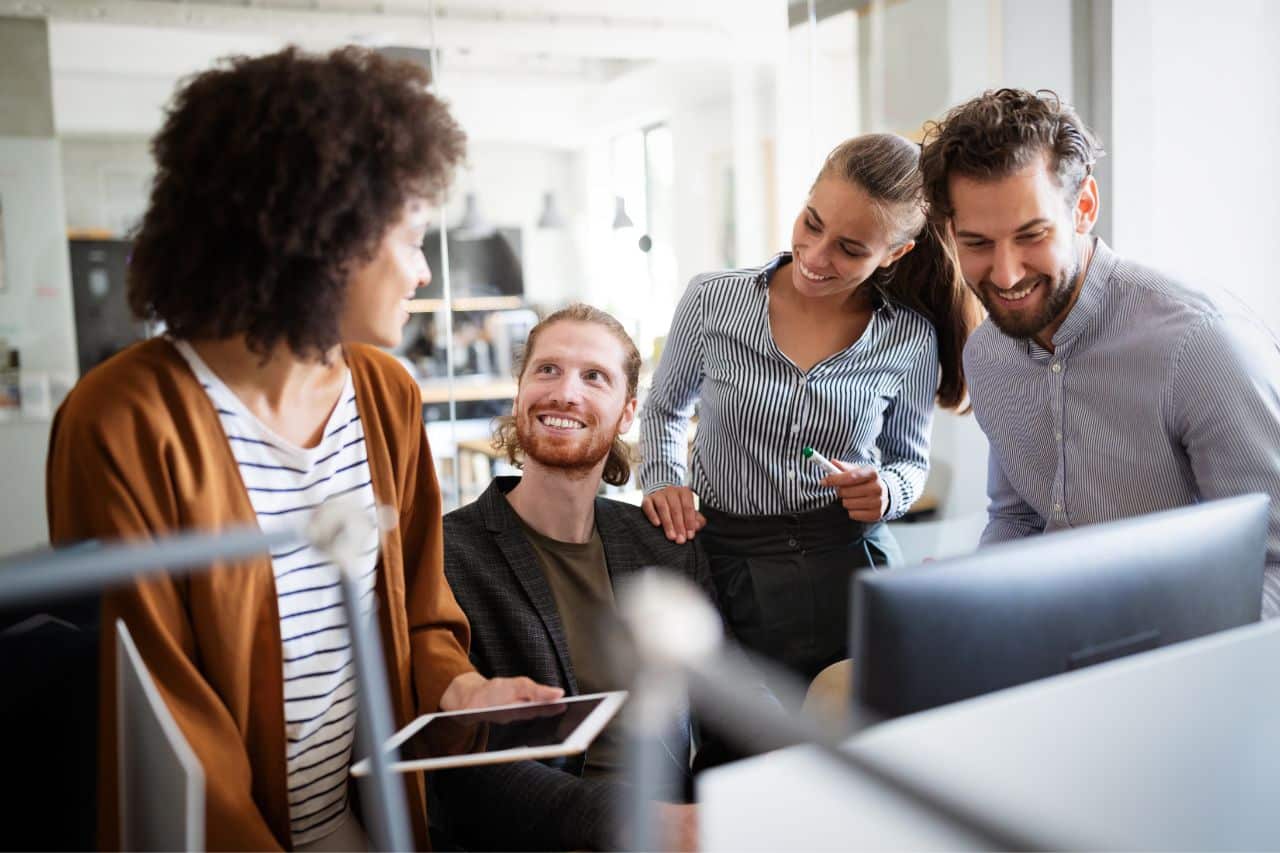 Das Gemälde zeigt eine Gruppe von vier lächelnden Menschen - zwei Frauen und zwei Männer, die sich um einen Computerbildschirm in einem Büro versammelt haben. Eine der Frauen mit lockigen, kurzen Haaren hält ein Tablet in der Hand, eine andere einen Marker. Einer der Männer sitzt auf einem Stuhl, der andere schaut auf den Computerbildschirm. Es scheint, als ob sie in eine Diskussion verwickelt sind oder zusammenarbeiten. Im Hintergrund sind Möbel und Büroausstattung zu sehen, was auf ein professionelles Umfeld hindeutet