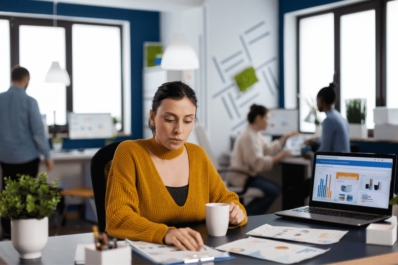 Das Foto zeigt ein Büro, in dem mehrere Personen an Schreibtischen arbeiten. Im Vordergrund sitzt eine Frau mit einem Laptop an einem Schreibtisch, auf dem verschiedene Grafiken angezeigt werden. Die Frau trägt einen orangefarbenen Pullover, ein Oberteil und hält eine weiße Tasse in der Hand. Auf dem Schreibtisch liegen Zettel mit Diagrammen, Büromaterial und eine Topfpflanze. Im Hintergrund sieht man andere Personen, die an Computern und großen Fenstern arbeiten, durch die natürliches Licht einfällt.