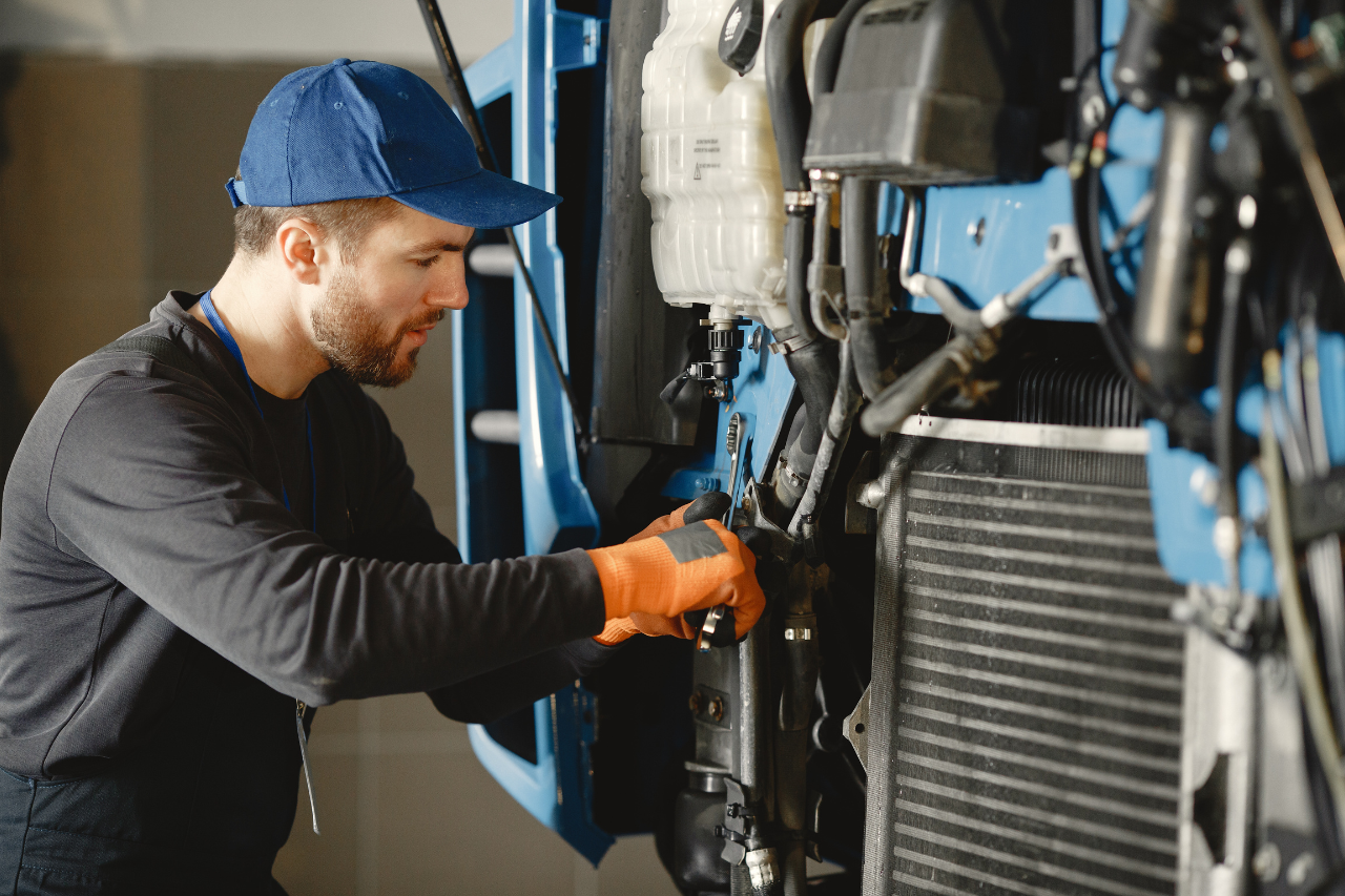 Das Gemälde zeigt eine Person mit blauem Hut und orangefarbenen Handschuhen, die an einer Maschine arbeitet. Die Person scheint Wartungs- oder Reparaturarbeiten an der Maschine durchzuführen, die verschiedene Komponenten wie den Kühler, die Schläuche und einen Tank für weiße Flüssigkeit umfasst.