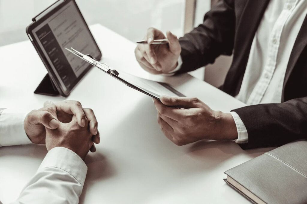 Na zdjęciu widać dwie osoby siedzące przy biurku podczas spotkania. Jedna z nich, ubrana w ciemną marynarkę i jasną koszulę, trzyma w dłoni długopis oraz clipboard z dokumentami. Naprzeciwko siedzi druga osoba w jasnej koszuli, z dłońmi splecionymi na blacie stołu. Na biurku znajduje się także tablet ustawiony na podstawce z wyświetlonym formularzem lub dokumentem oraz zamknięty notes.