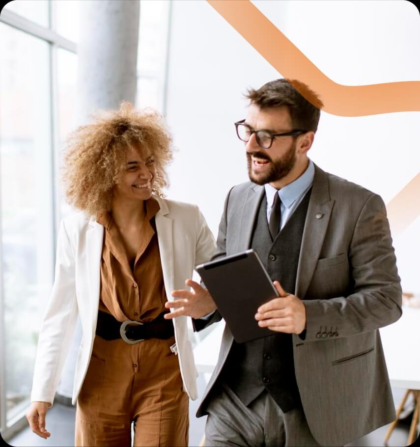 Na zdjęciu widać dwoje profesjonalnie ubranych osób idących razem przez jasne, nowoczesne wnętrze biurowe z dużymi oknami. Kobieta ma kręcone, jasnobrązowe włosy i ubrana jest w brązowy kombinezon z paskiem oraz jasną marynarkę. Uśmiecha się i patrzy na swojego towarzysza. Mężczyzna ma na sobie elegancki, szary garnitur z kamizelką i krawatem oraz okulary. W ręku trzyma tablet i gestykuluje podczas rozmowy. 