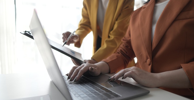 Na zdjęciu widać dwie osoby pracujące przy biurku w jasnym, nowoczesnym wnętrzu biurowym. Na pierwszym planie znajduje się otwarty laptop, na którego klawiaturze jedna z osób pisze. Ubrana jest w elegancką, brązową marynarkę, co sugeruje formalny, biznesowy charakter spotkania.