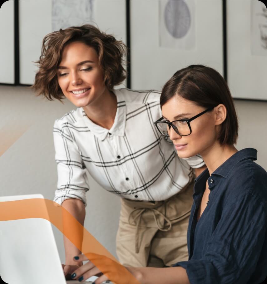 Na zdjęciu widać dwie kobiety pracujące wspólnie przy laptopie w jasnym, nowoczesnym wnętrzu biurowym. Jedna z nich siedzi przed komputerem, ma krótkie, ciemne włosy i okulary. Skupiona patrzy na ekran, trzymając dłoń na klawiaturze. Ubrana jest w ciemną koszulę, co nadaje jej profesjonalny wygląd. Druga uśmiechnięta kobieta stoi obok i lekko pochyla się nad laptopem. Ma krótkie, falowane włosy i jasną koszulę w kratę.