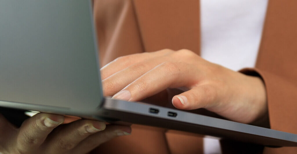 Na zdjęciu widoczne jest zbliżenie dłoni osoby korzystającej z laptopa. Fotografia skupia się na pracy przy komputerze i podkreśla wykorzystanie nowoczesnych technologii w codziennych obowiązkach zawodowych.